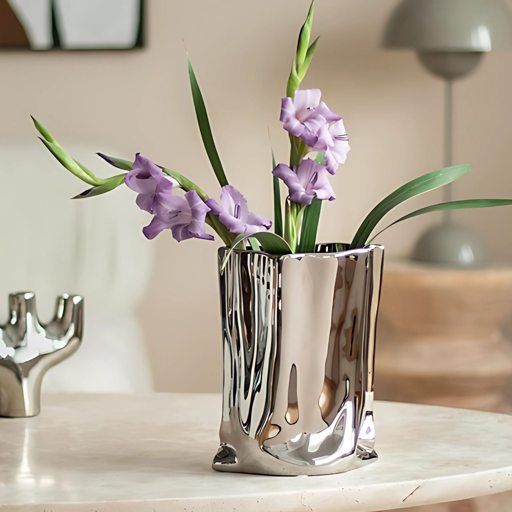 Silver, chrome bag vase on tabletop with purple flowers.