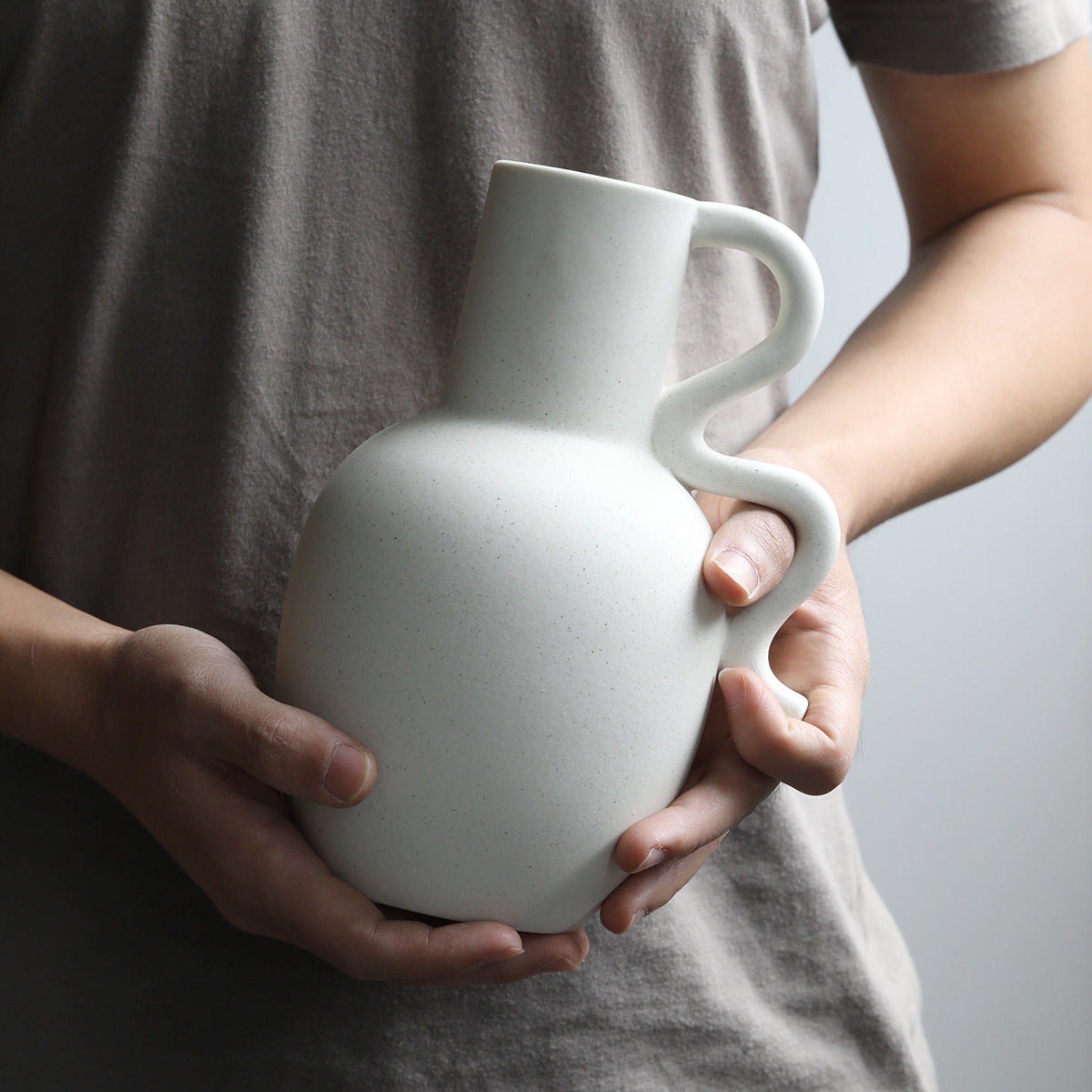 Person holding a white, ceramic, pitcher vase with wavy handle.