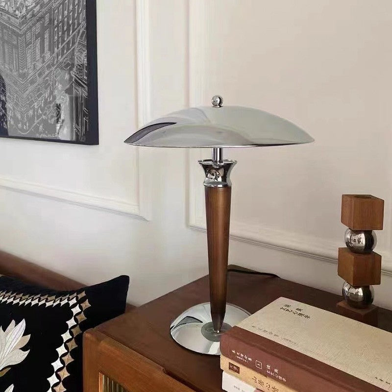 A table lamp with a wooden pole and a silver chrome base and lamp shade on top of a dresser with books and candleholder in living room.