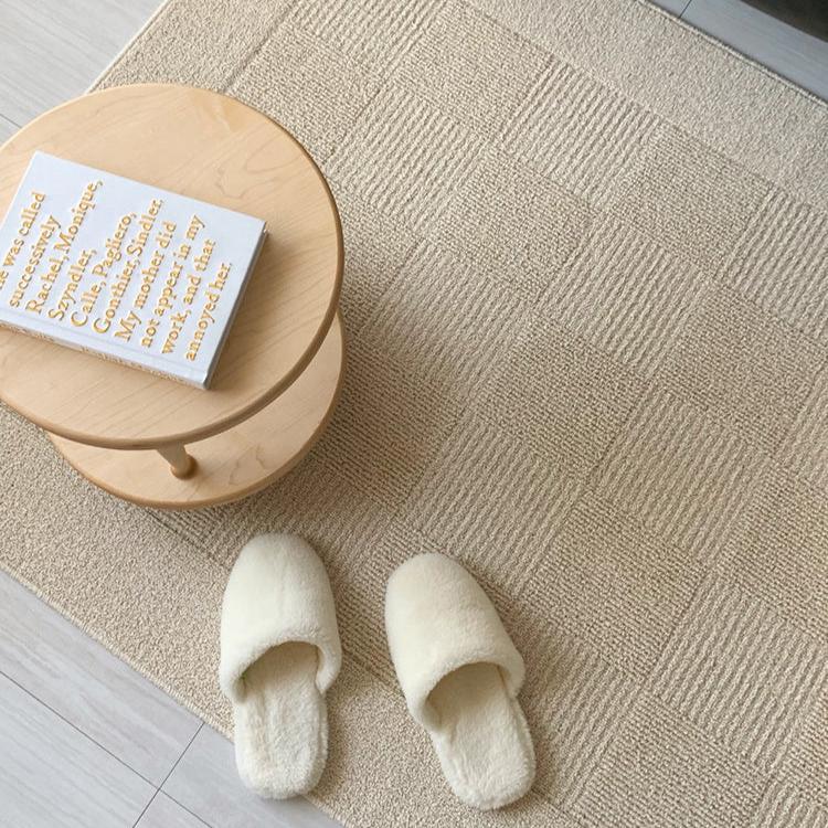 A beige floor rug with a gridded texture and a small table and pair of slippers.