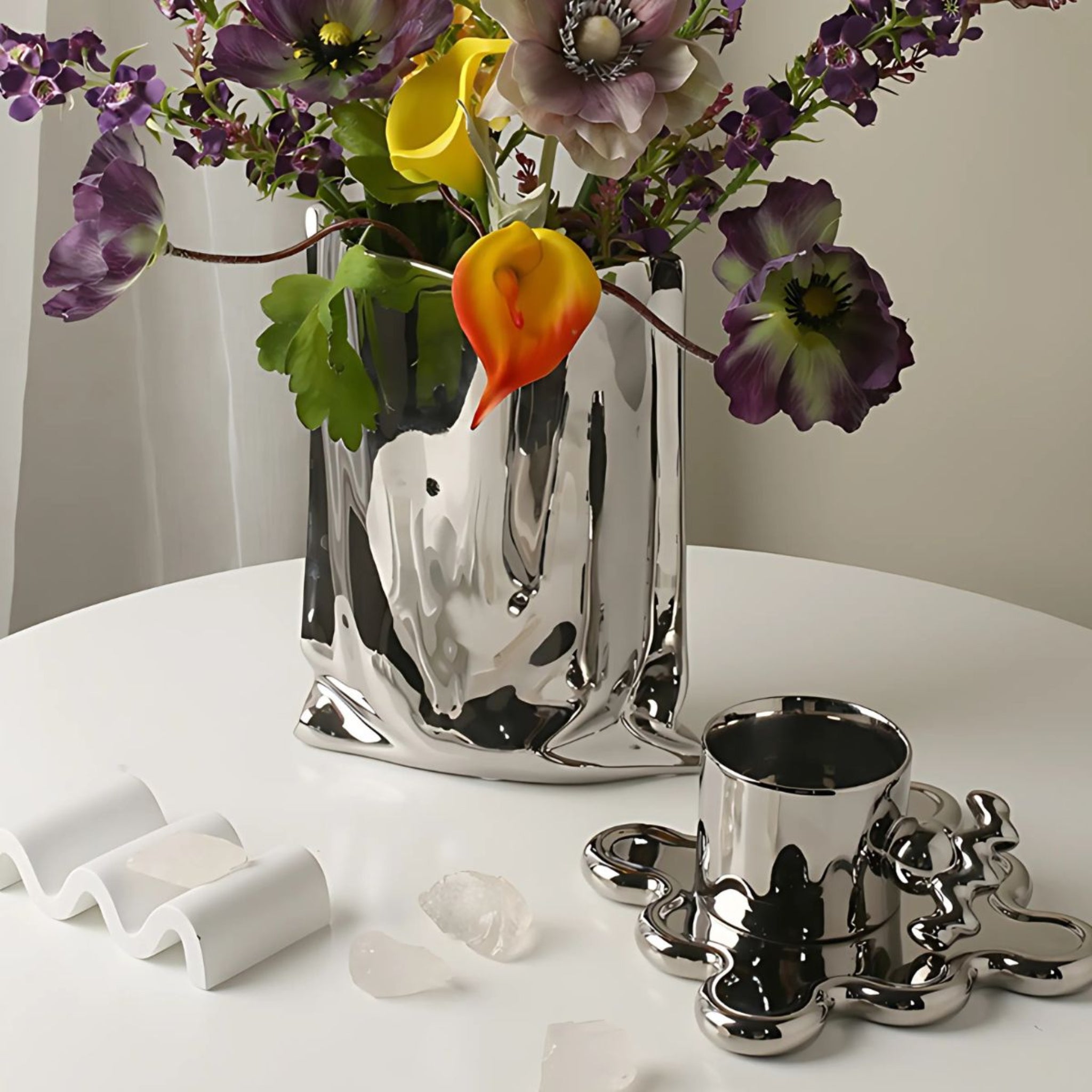 A table with electroplated silver chrome home decor including a vase and a coffee mug with saucer.
