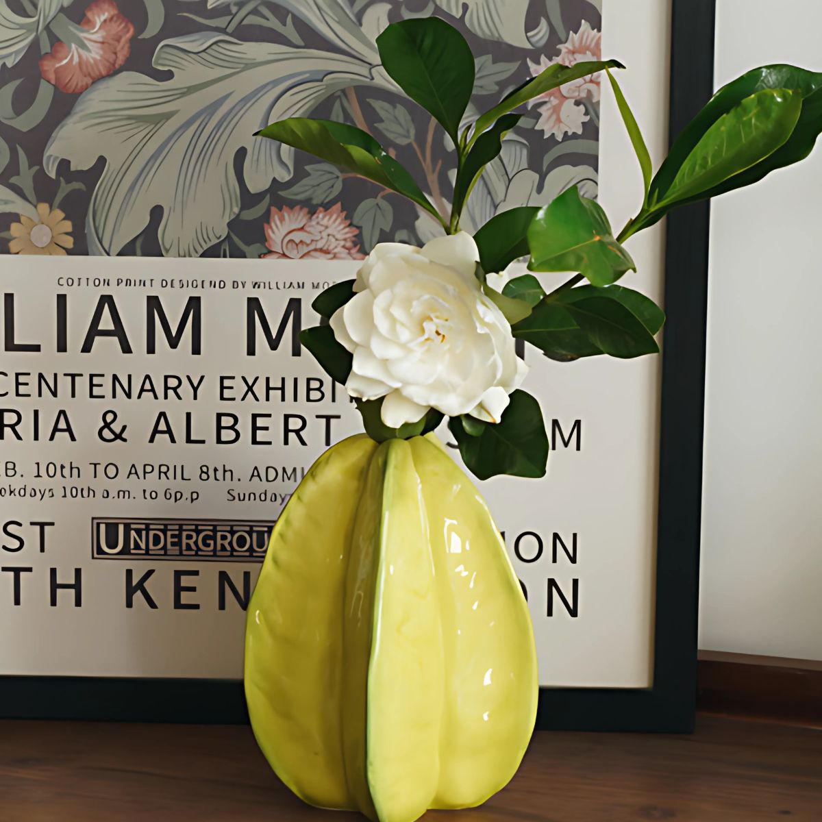 Wooden table with William Morris poster and yellow, ceramic, starfruit design vase holding green leafs and white flower.