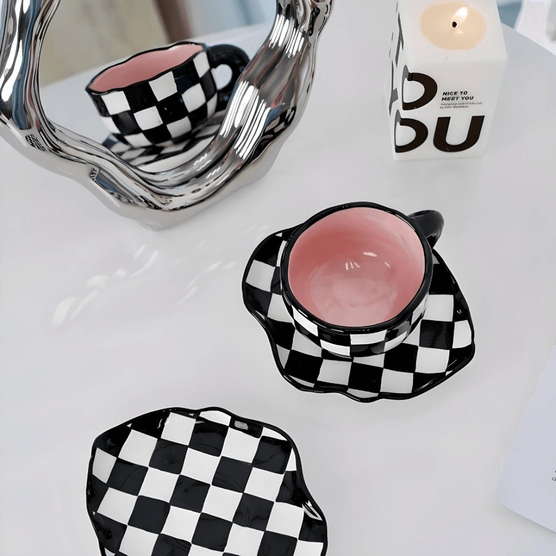Black white pink checkerboard irregular ceramic mug and saucer on table with mirror and candle